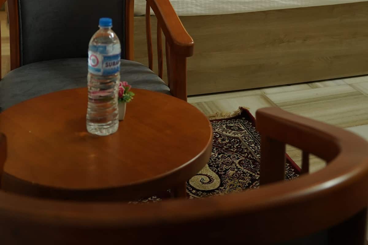 Quarto de hotel com uma cama, uma mesa de cabeceira, uma cadeira, uma mesa com uma garrafa de água e um ventilador de teto.