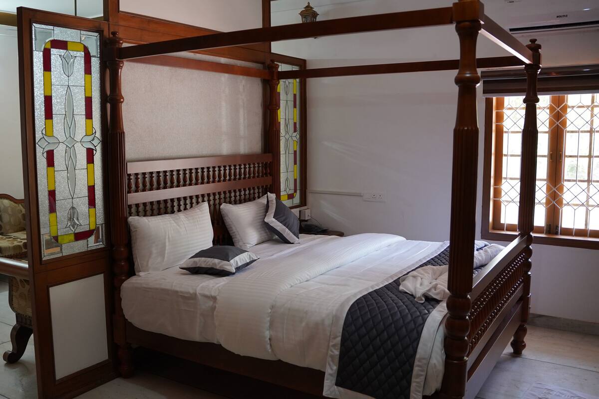 Uma cama de dossel com estrutura de madeira e colcha branca, um banco com almofada estampada e uma janela com grade decorativa.
