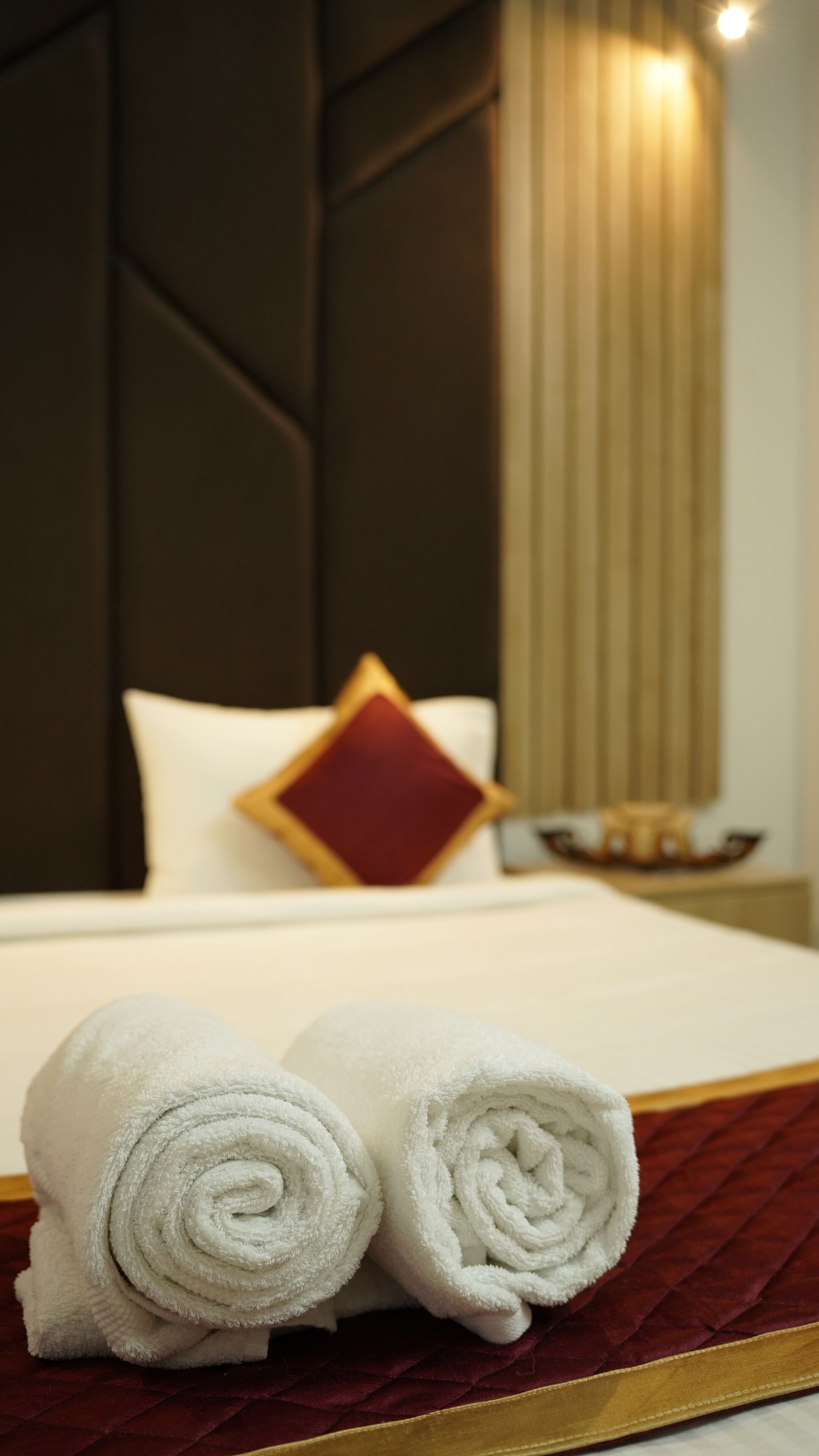 A neatly made bed with white linens and a red accent pillow, a rolled white towel, and a dark headboard.
