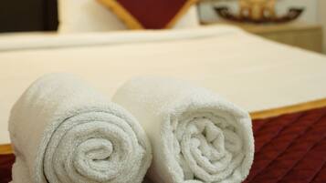 A neatly made bed with white linens and a red accent pillow, a rolled white towel, and a dark headboard.