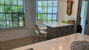 A kitchen with a large island, two windows with blinds, a clock on the wall, and a door with a window. - Fabulous Naples Townhouse in Desirable Pelican Ridge Location (Naples)