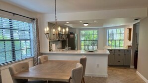 A modern kitchen with a central island, a dining area with a large table and chairs, and large windows with blinds. - Fabulous Naples Townhouse in Desirable Pelican Ridge Location (Naples)