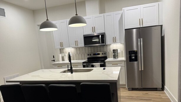 A modern kitchen with white cabinets, a stainless steel refrigerator, a built-in microwave, and a bar area with stools. - Elegant Plush  Properties (Houston)