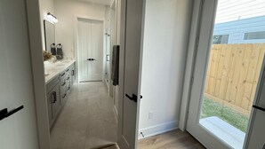 A hallway leading to a bathroom with a shower, a sink, and a mirror. - Elegant Plush  Properties (Houston)