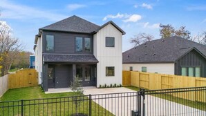 A two-story residential building with a grey roof, a white lower section, and a visible front yard with a wooden fence and a black metal gate. - Elegant Plush  Properties (Houston)
