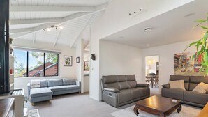 A spacious living room with a grey sectional sofa, a dark brown leather sofa, and a wooden coffee table. There is a large window providing natural light and a view of the outside. - Waiake Family Retreat - Auckland Holiday Home (Auckland)