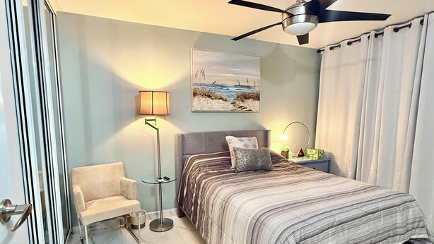 Een slaapkamer met een bed, een stoel, een plafondventilator, een schilderij en een vloerlamp.