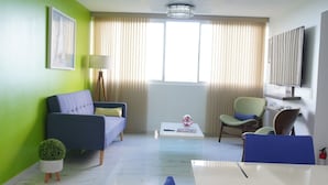 Een moderne woonkamer met een blauwe bank, een eettafel met stoelen, een kleine potplant en een kroonluchter.
