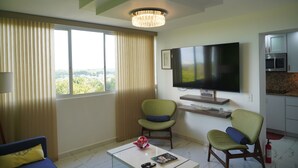 Een moderne woonkamer met een grote televisie, twee groene stoelen, een salontafel met een boek en afstandsbedieningen, en uitzicht op groen door het raam.