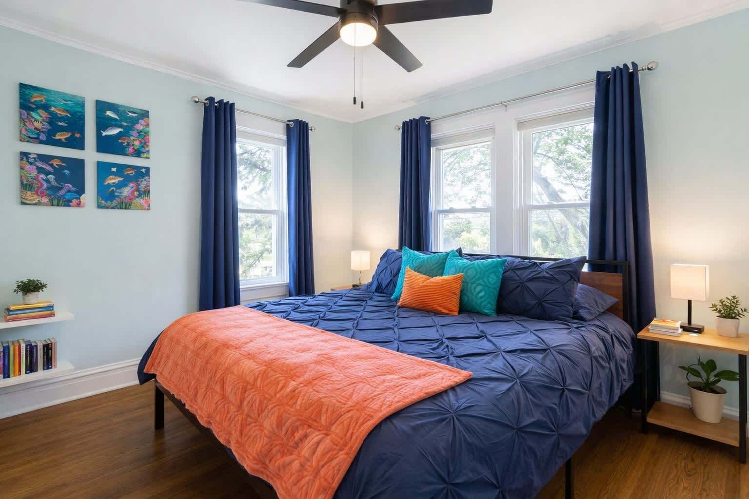 Ein Schlafzimmer mit einem Bett, zwei Fenstern mit Vorhängen, einem Deckenventilator und einem Kunstwerk an der Wand.