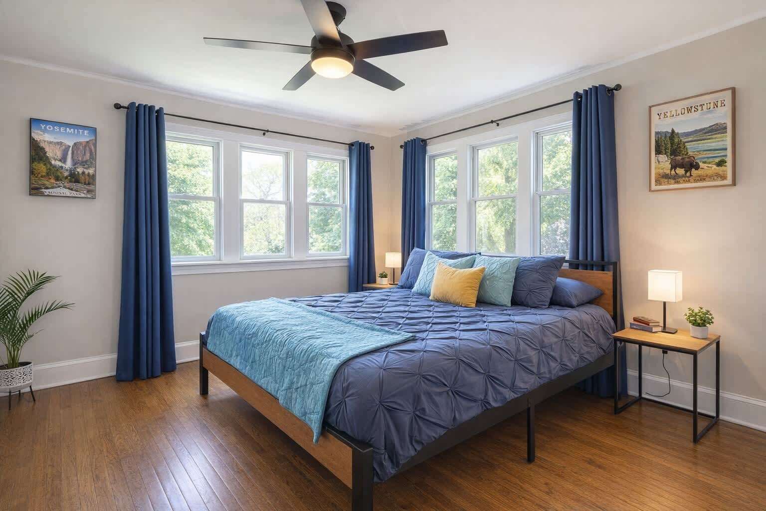 Une chambre à coucher avec un lit, un ventilateur de plafond, une table de chevet, une lampe et une fenêtre avec des rideaux.