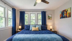 Un dormitorio con una cama grande, dos mesitas de noche, un ventilador de techo y una ventana con cortinas.