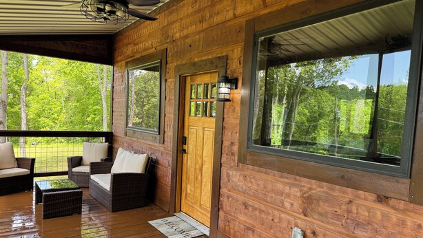 Een houten blokhut met een veranda, rieten meubilair en grote ramen met uitzicht op een bos.