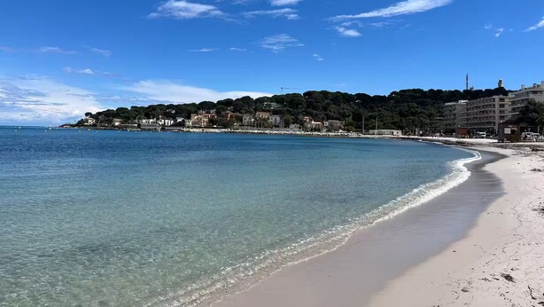 Une plage côtière aux eaux cristallines, avec un rivage sablonneux et des bâtiments visibles à l’arrière-plan.