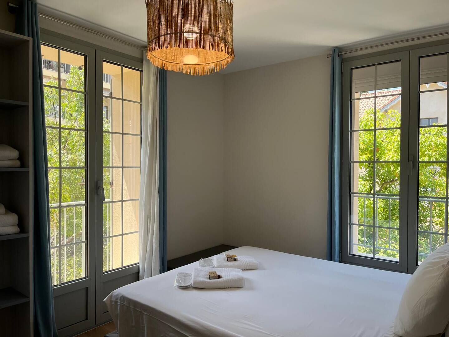A hotel room with a bed, two windows with curtains, a shelf with towels, and a hanging light fixture.