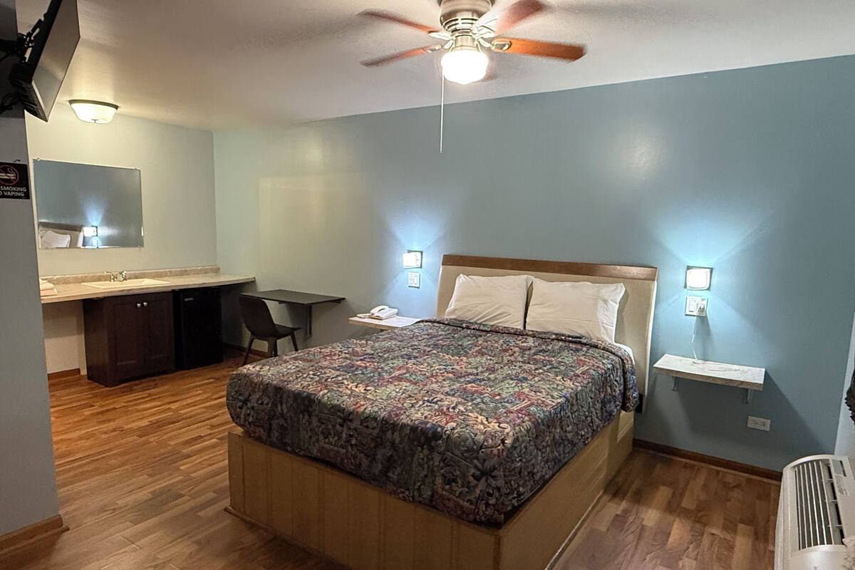 A bedroom with a bed, a desk, a chair, a mirror, and a ceiling fan.