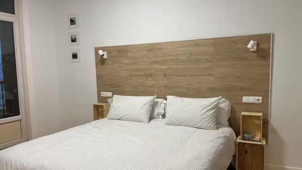 Een net opgemaakt bed met een houten hoofdbord, twee nachtkastjes met lampen en een raam met jaloezieën.