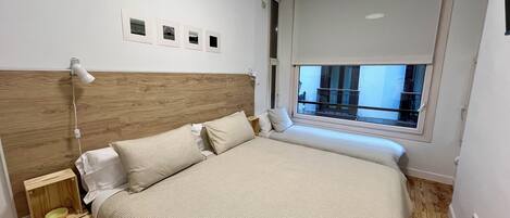 A bedroom with a wooden headboard, a bed with beige bedding, a bedside table with a lamp, and a window with a view of the outside.