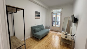 A room with a wooden floor, a green sofa, a small table with a mirror, and a TV mounted on the wall. - Pensión Miramar (San Sebastian)
