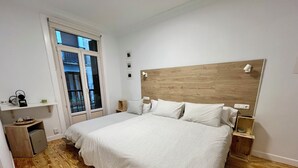 A bedroom with a large bed, a wooden headboard, a small bedside table, a nightstand, and a window with blinds. - Pensión Miramar (San Sebastian)