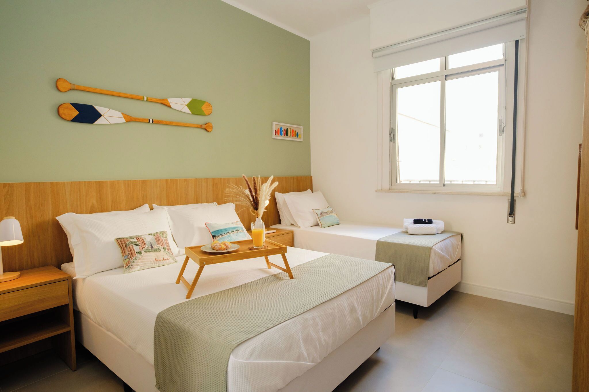 Une chambre d’hôtel avec deux lits, une tête de lit en bois, une table de chevet avec une lampe, une fenêtre et une planche de paddle fixée au mur.