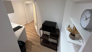 A narrow hallway with a wooden floor, a white door at the end, and a black shelf unit with a clock on the wall. - Large American Studio (Toulon)