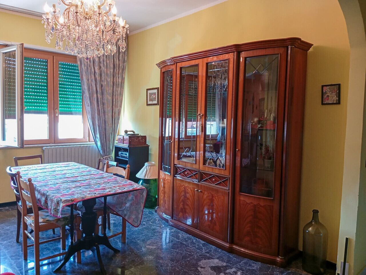 A dining room with a wooden cabinet, a table with chairs, a chandelier, and a window with blinds.