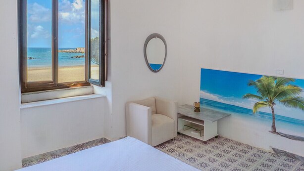 Ein Zimmer mit großem Fenster und Blick auf einen Strand, ein kleines weißes Sofa, ein Nachttisch mit Lampe und ein Wandbild einer Palme.