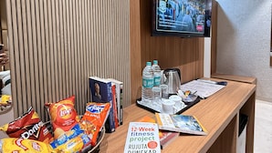 A reception desk with snacks, a book titled "The 12-Week fitness project," and a television displaying a news program. - Hotel Akaay (Ujjain)