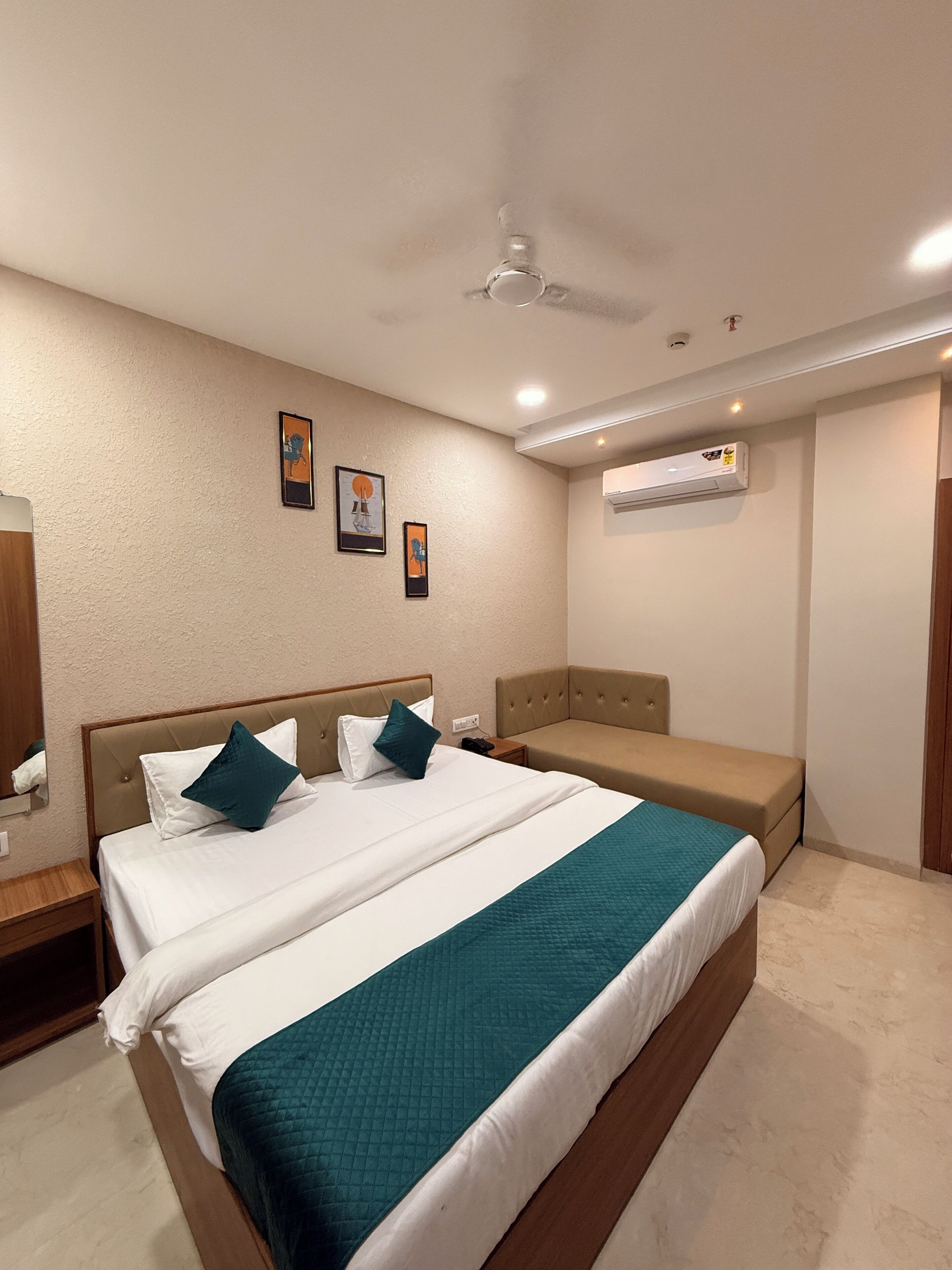 A hotel room with a large bed, a bedside table, a nightstand, a ceiling fan, an air conditioning unit, and two framed pictures on the wall.