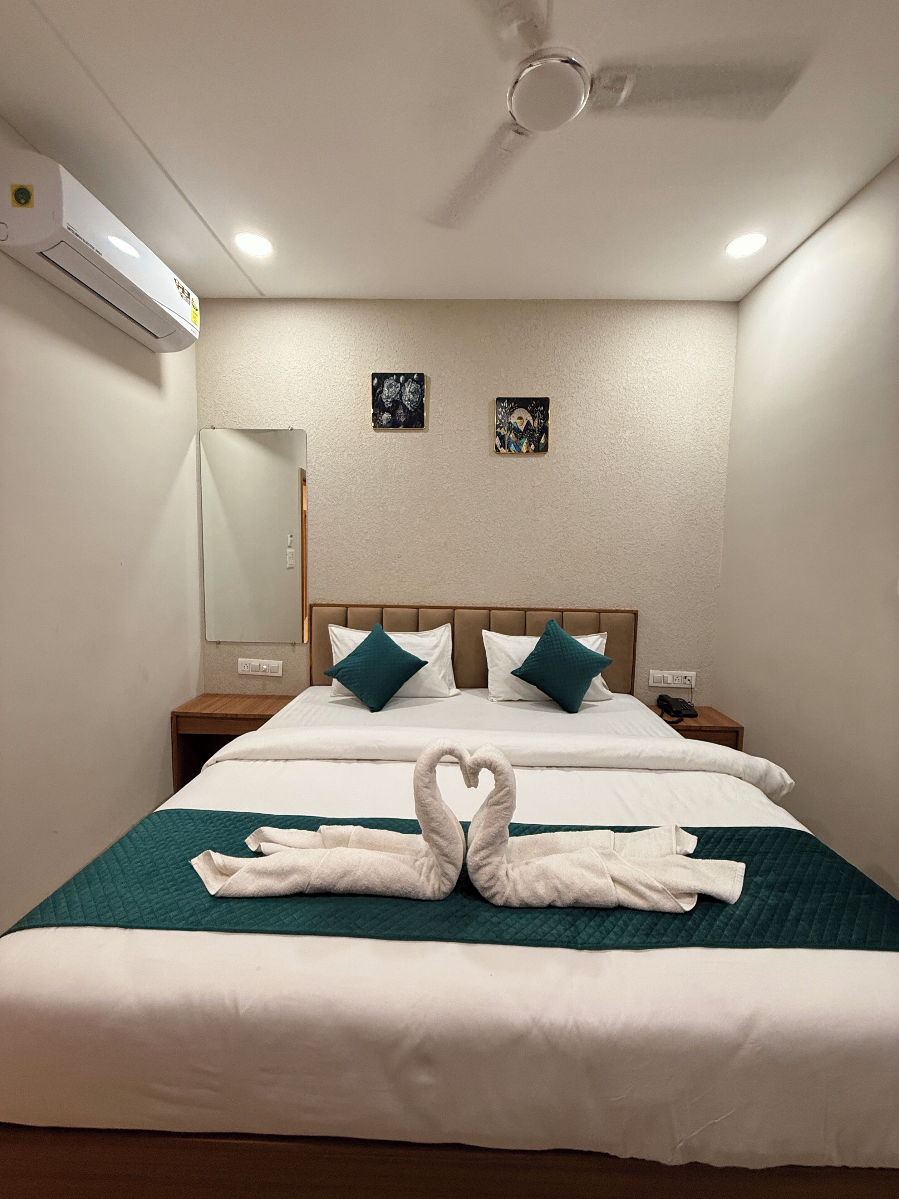 A hotel room with a bed, two pillows, a bedside table, a nightstand, a mirror, and a wall-mounted air conditioner.