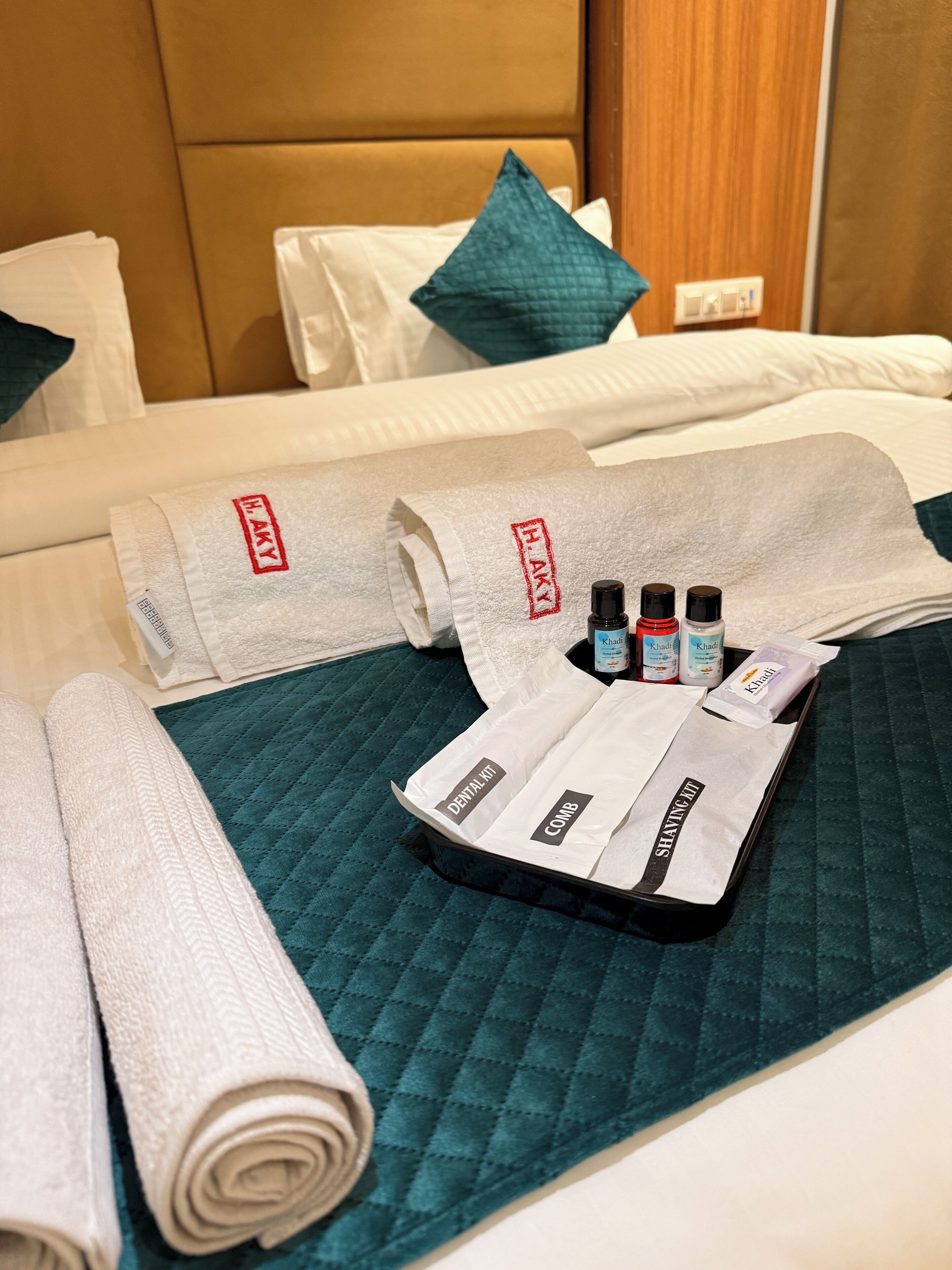 A neatly made bed with a teal quilted bedspread, white pillows, and a rolled white towel. A tray with a comb, shaving kit, and three bottles of hair product is placed on the bed.