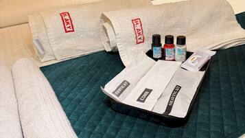 A neatly made bed with a teal quilted bedspread, white pillows, and a rolled white towel. A tray with a comb, shaving kit, and three bottles of hair product is placed on the bed.