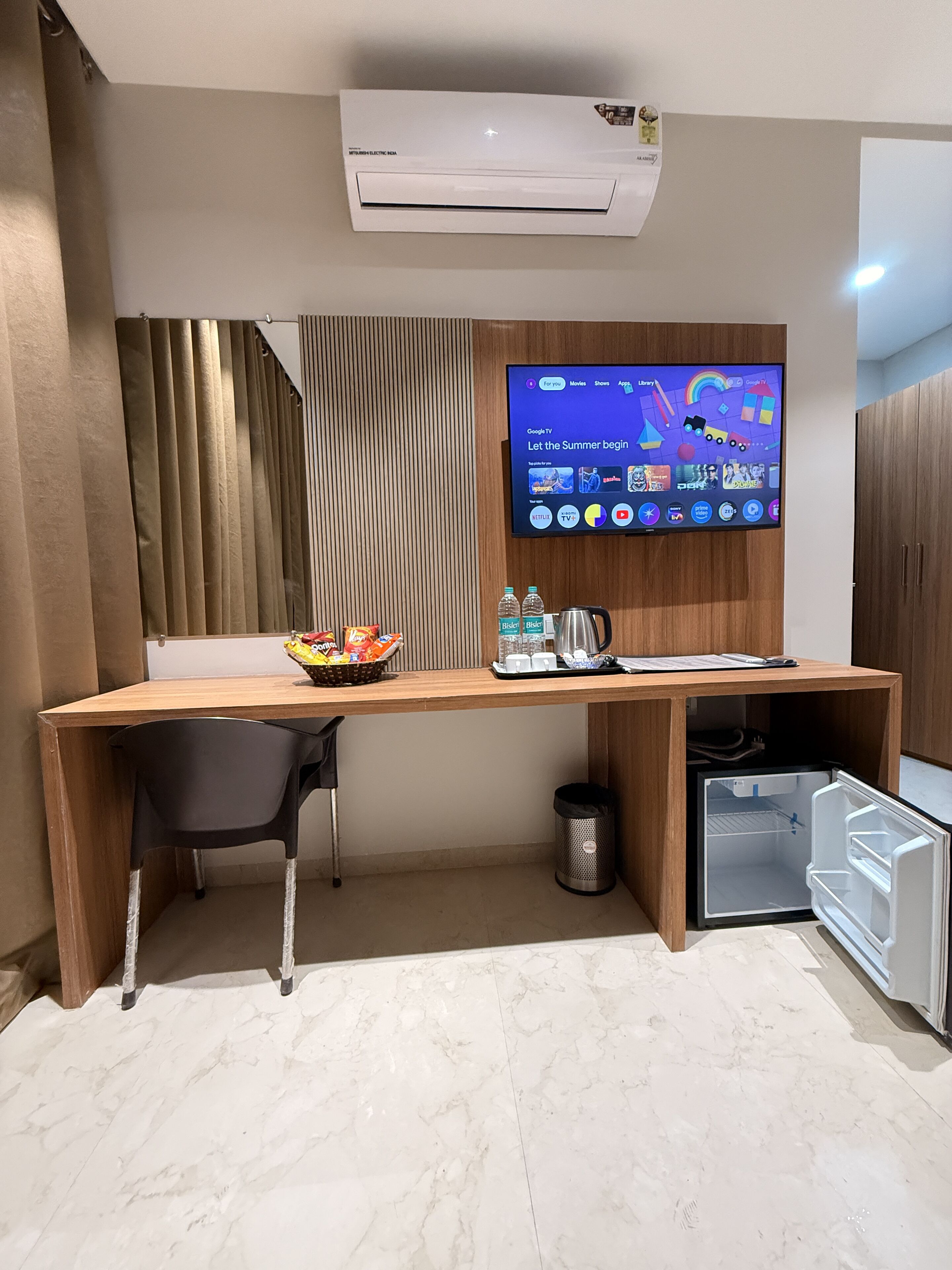 Habitación de hotel con un escritorio de madera, una silla negra, un televisor montado en la pared y un minibar.