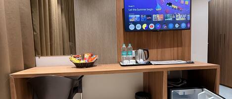 Habitación de hotel con un escritorio de madera, una silla negra, un televisor montado en la pared y un minibar.
