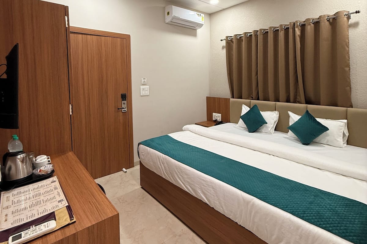 A hotel room with a bed, a wooden wardrobe, a desk with a kettle and a menu, and an air conditioning unit.