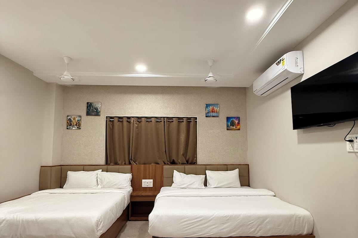 A hotel room with two single beds, a ceiling fan, an air conditioning unit, a flat-screen TV mounted on the wall, and artwork on the walls.