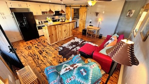Una sala de estar acogedora con suelo de madera, un sofá con cojines estampados, una silla roja y un comedor con mesa y sillas.