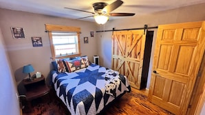 Un dormitorio con cama, mesita de noche, lámpara, ventilador de techo, ventana y puerta de madera.