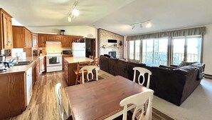En rummelig opholdsstue med et træbord og stole, en mørk sofa og et køkken med træskabe og moderne hårde hvidevarer.
