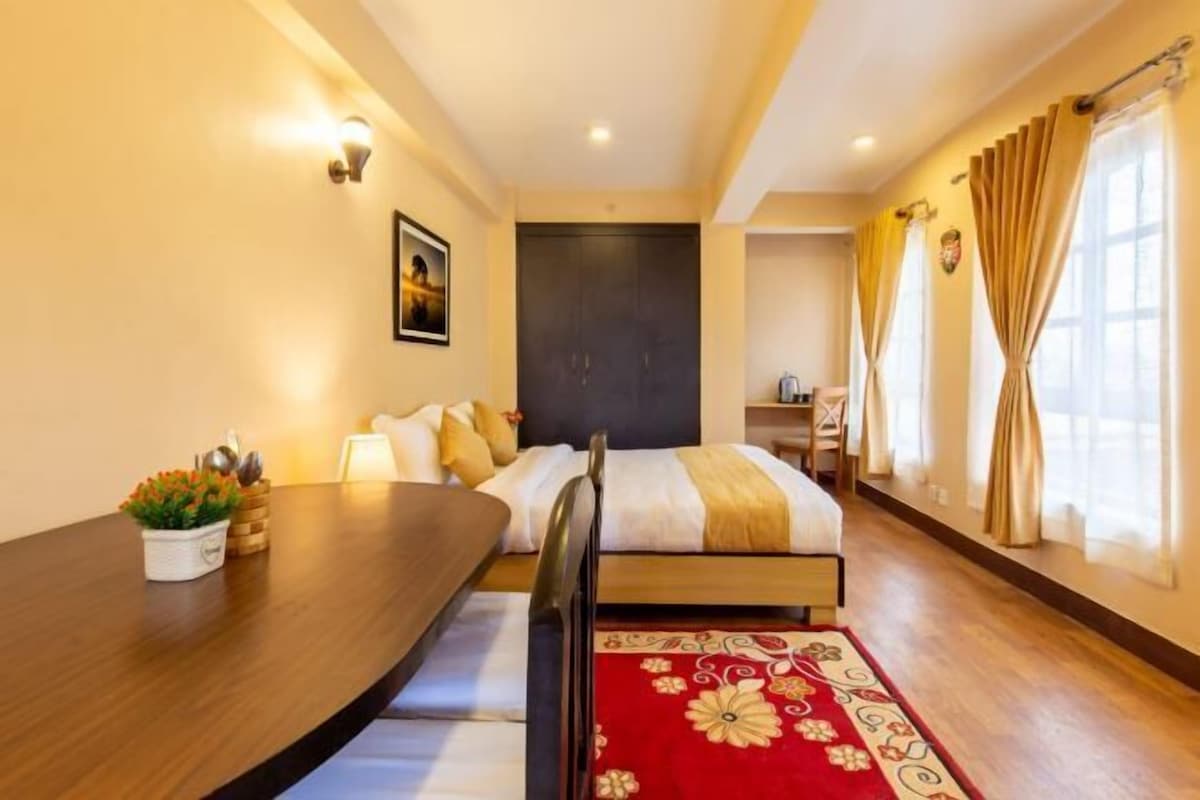 A hotel room with a bed, a desk, a chair, a red patterned rug, and a wall-mounted picture.