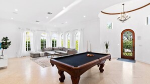 A spacious living room with a pool table, a sofa set, and large windows.