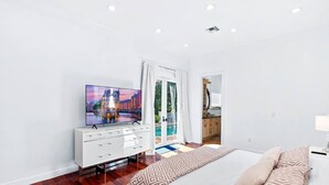 A modern bedroom with a TV, a bed with patterned pillows, a wooden floor, and a view of an outdoor pool area.