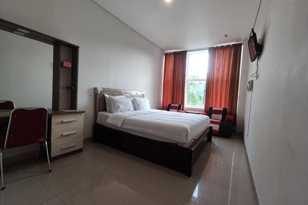 Un dormitorio con cama, escritorio, silla, televisor y ventana con cortinas.