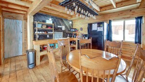 Interior de una cabaña de madera con una mesa de comedor, sillas, una zona de cocina con electrodomésticos y un ventanal grande.