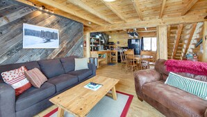 Un acogedor interior de cabaña con techo de madera, un sofá gris, un sofá de cuero marrón, una mesa de centro de madera y una zona de comedor con mesa y sillas de madera.