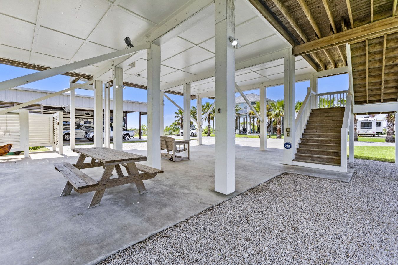 An outdoor covered patio area with picnic tables, a staircase, and a view of parked vehicles and houses in the background.