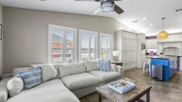 A living room with a sofa, coffee table, and a ceiling fan. There is a kitchen area with white cabinets and a blue island.