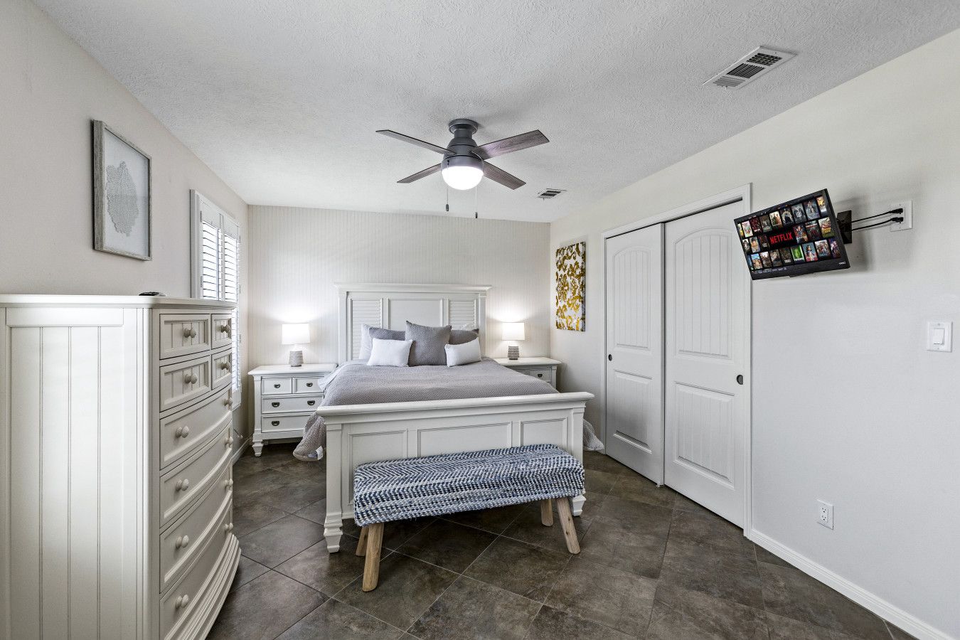 A bedroom with a bed, bedside tables, a bench, a ceiling fan, a wardrobe, and a wall-mounted TV.
