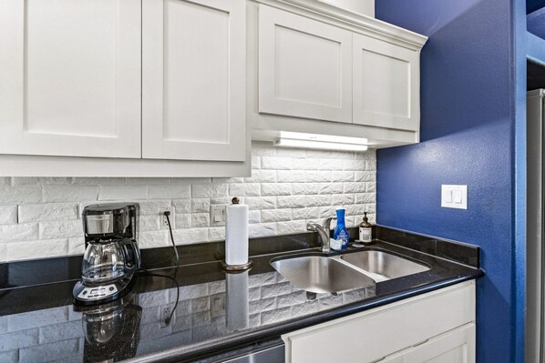 A kitchen with white cabinets, a black countertop, a coffee maker, a paper towel holder, and a sink with a faucet.